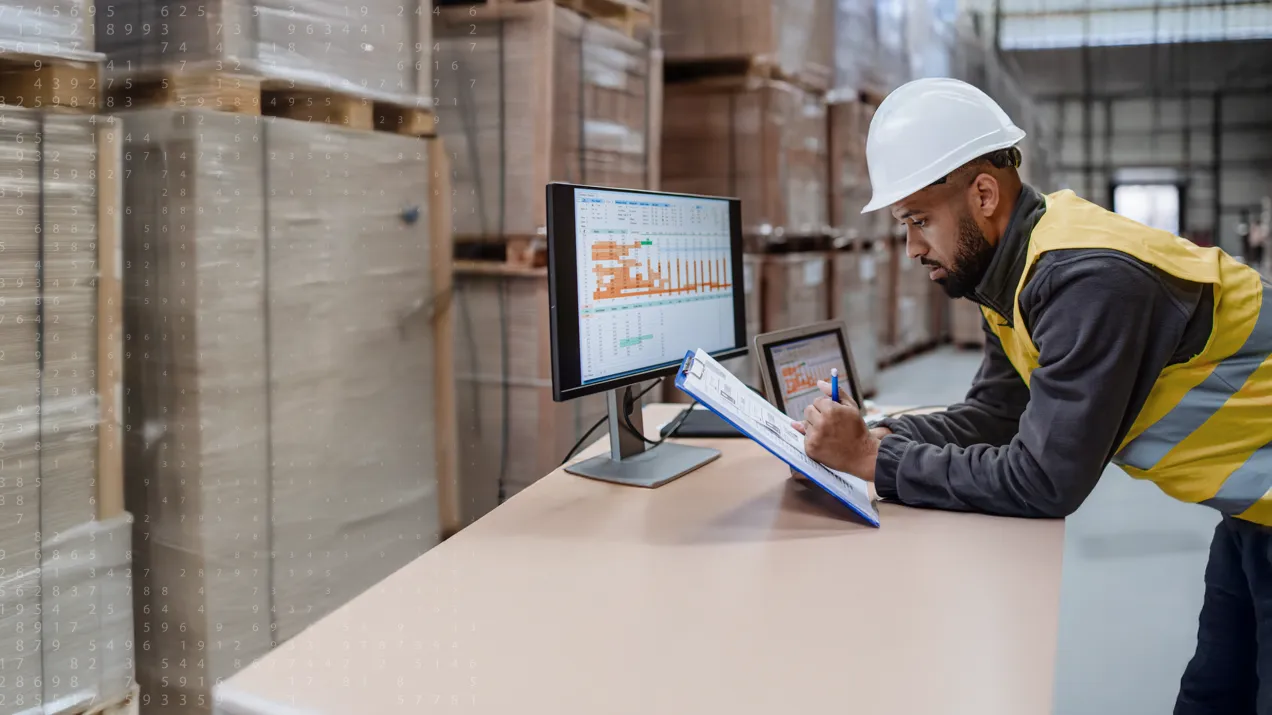 A construction worker holding a clipboard over a desk with a computer.