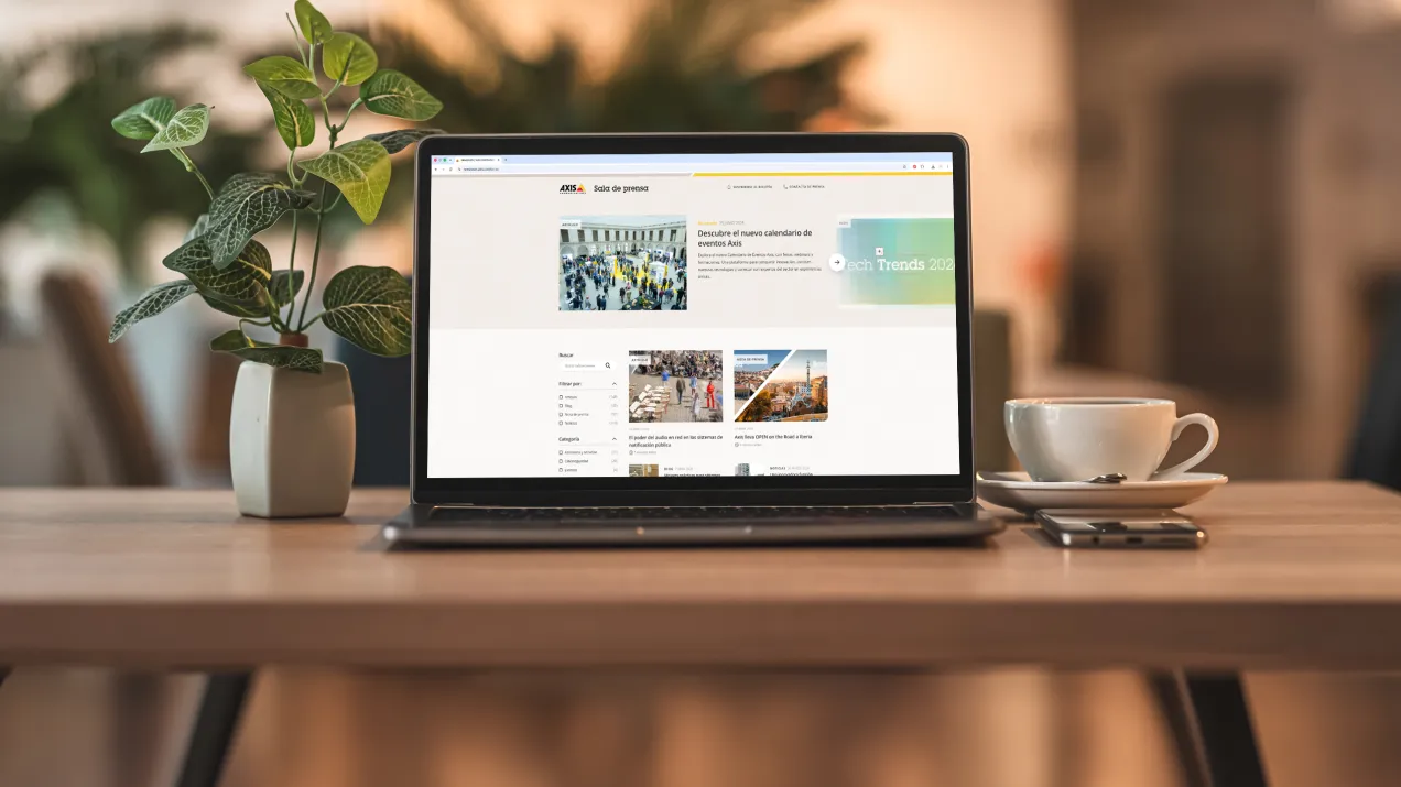 Laptop screen displaying the Axis Communications newsroom in Spanish, with a potted plant and a cup of coffee in the background.
