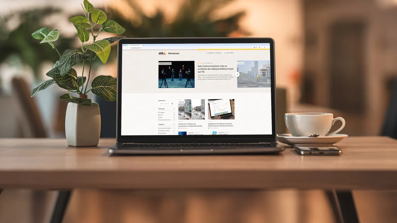 Laptop screen displaying the Axis Communications newsroom in french, with a potted plant and a cup of coffee in the background.