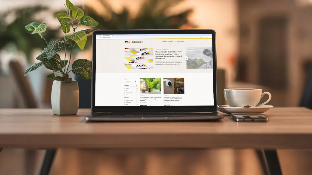 Laptop screen displaying the Axis Communications newsroom in Italian, with a potted plant and a cup of coffee in the background.