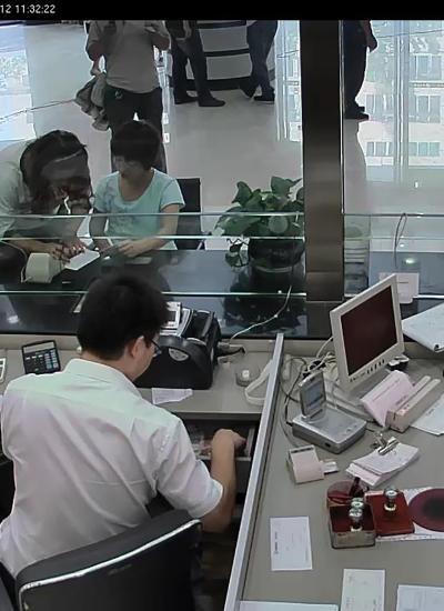 Two people at bank behind glass window assisting people.