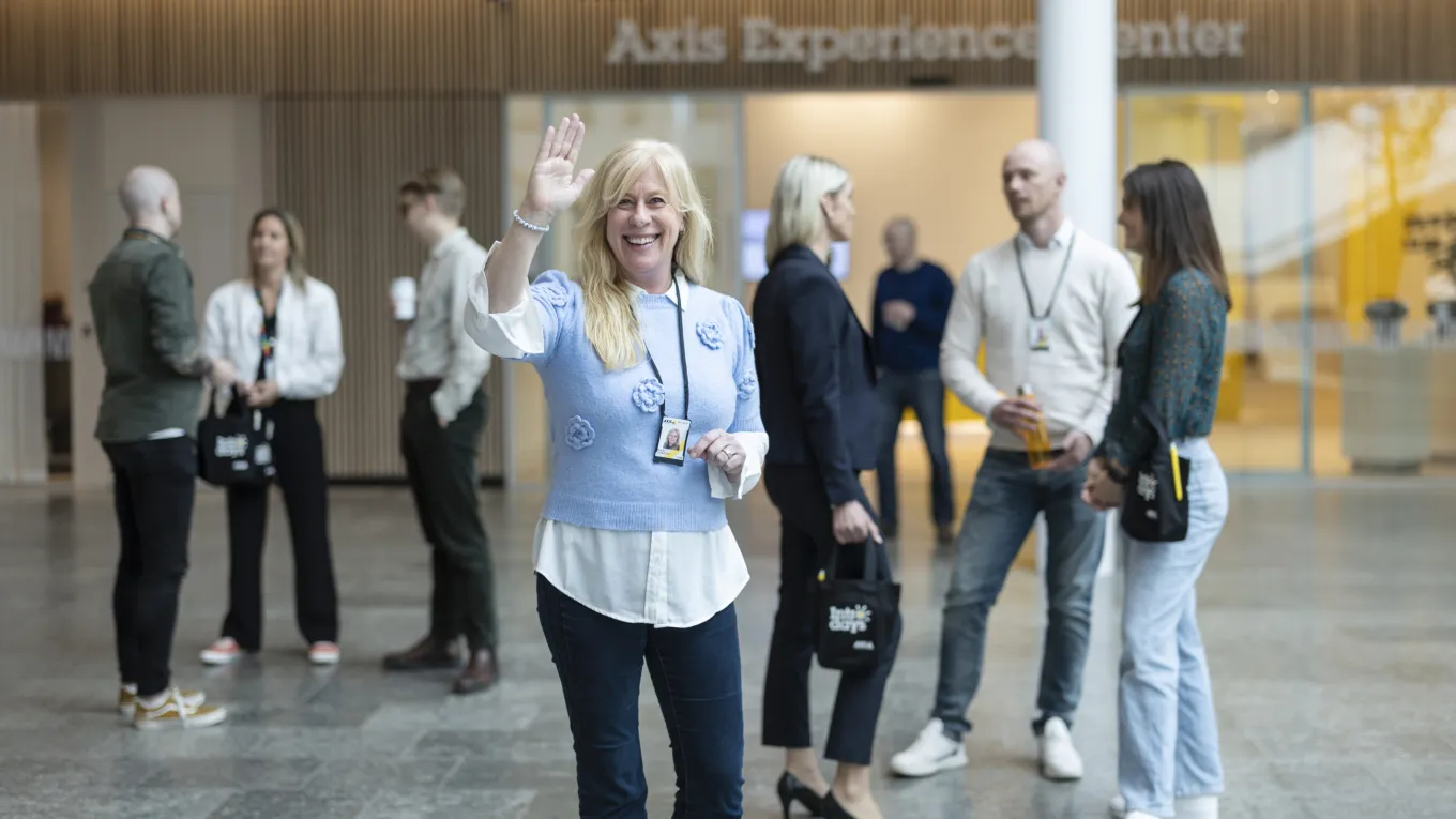 Malin Svensson standing in front of the camera, smiling and waving, with people standing in the background talking.