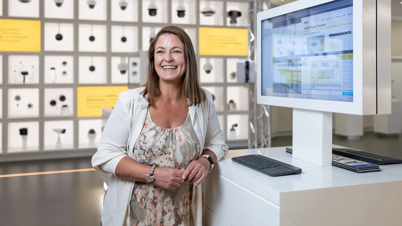 Vanessa Tyne, Strategic Customers Technical Manager at Axis, standing infront of the product wall in Axis Experience Center, smiling. 