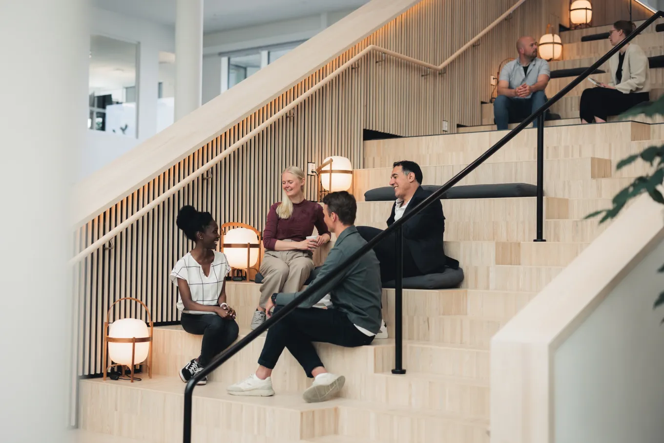 A group of Axis colleagues having a casual conversation and smiling while sitting on the large wooden stairs at the headquarters.