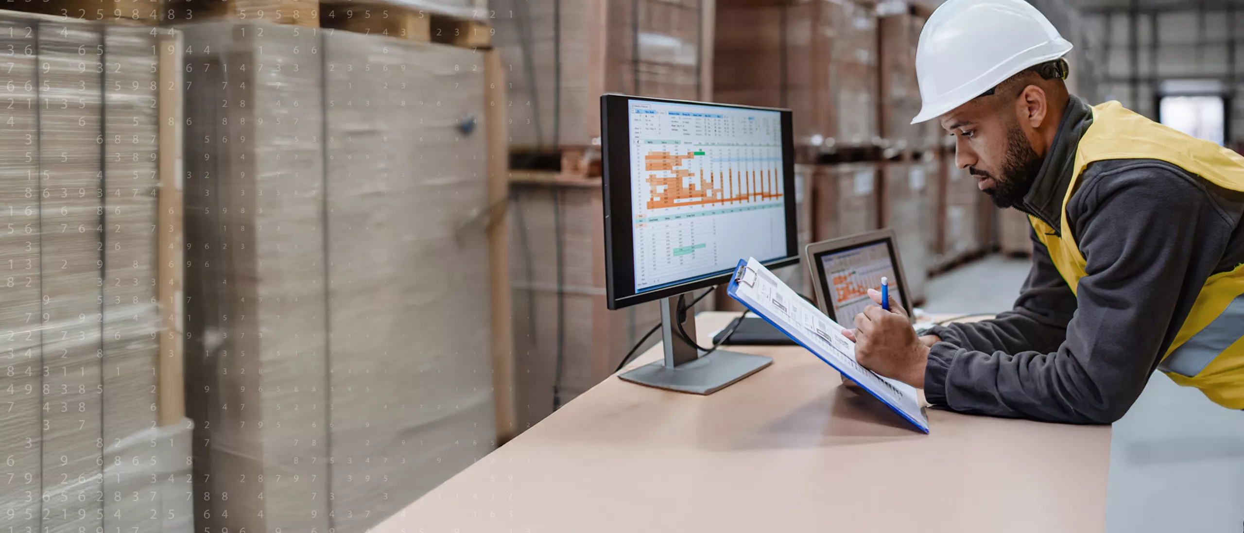A construction worker holding a clipboard over a desk with a computer.