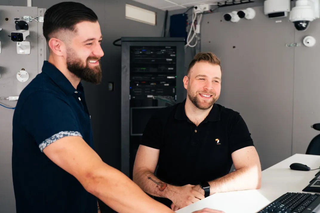 Two smiling men looking at a computer screen. Several Axis cameras hanging on the walls behind them.